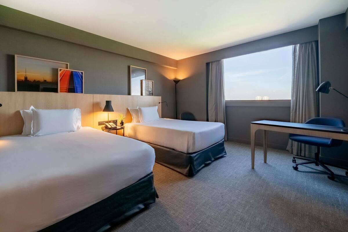 Habitación de hotel con dos camas, un escritorio, una silla, una lámpara y vistas a la ciudad.