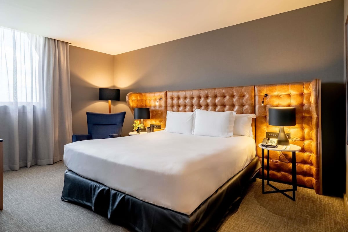 Una habitación de hotel con una cama grande, dos mesitas de noche con lámparas, una silla y una ventana con cortinas.