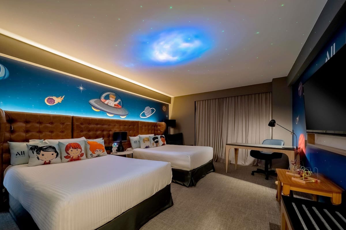 Habitación de hotel con dos camas, un escritorio, una silla, un televisor y una proyección de la galaxia en el techo.