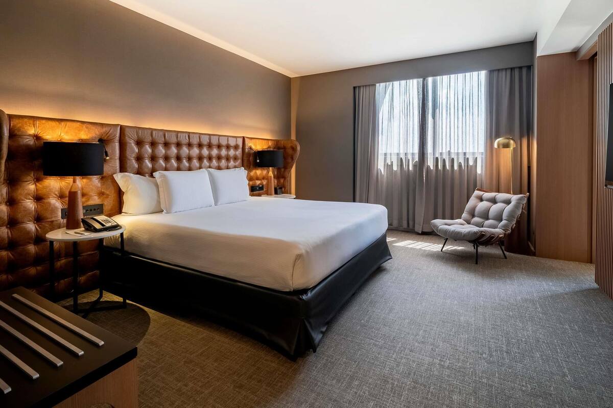 Habitación de hotel con una cama grande, una mesita de noche con lámpara, una silla y vistas al exterior a través de la ventana.