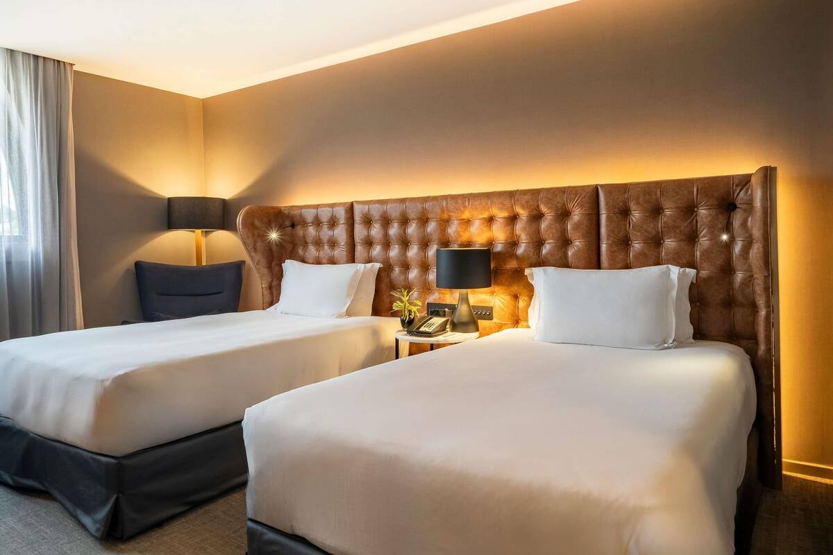 Habitación de hotel con dos camas, un cabecero grande y capitonado, lámparas de noche y un teléfono en la mesita.