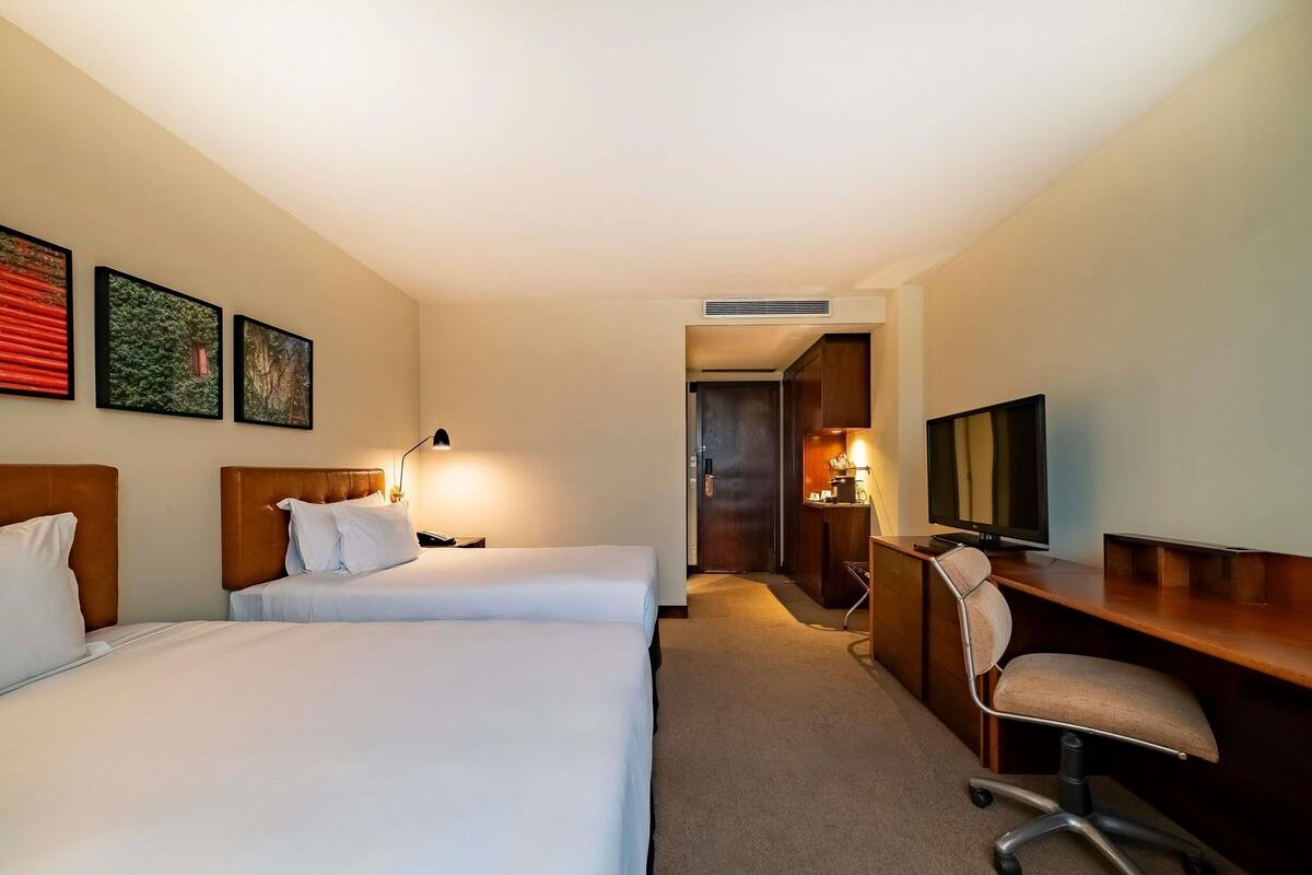 Habitación de hotel con dos camas, un escritorio, una silla, un televisor y obras de arte en la pared.
