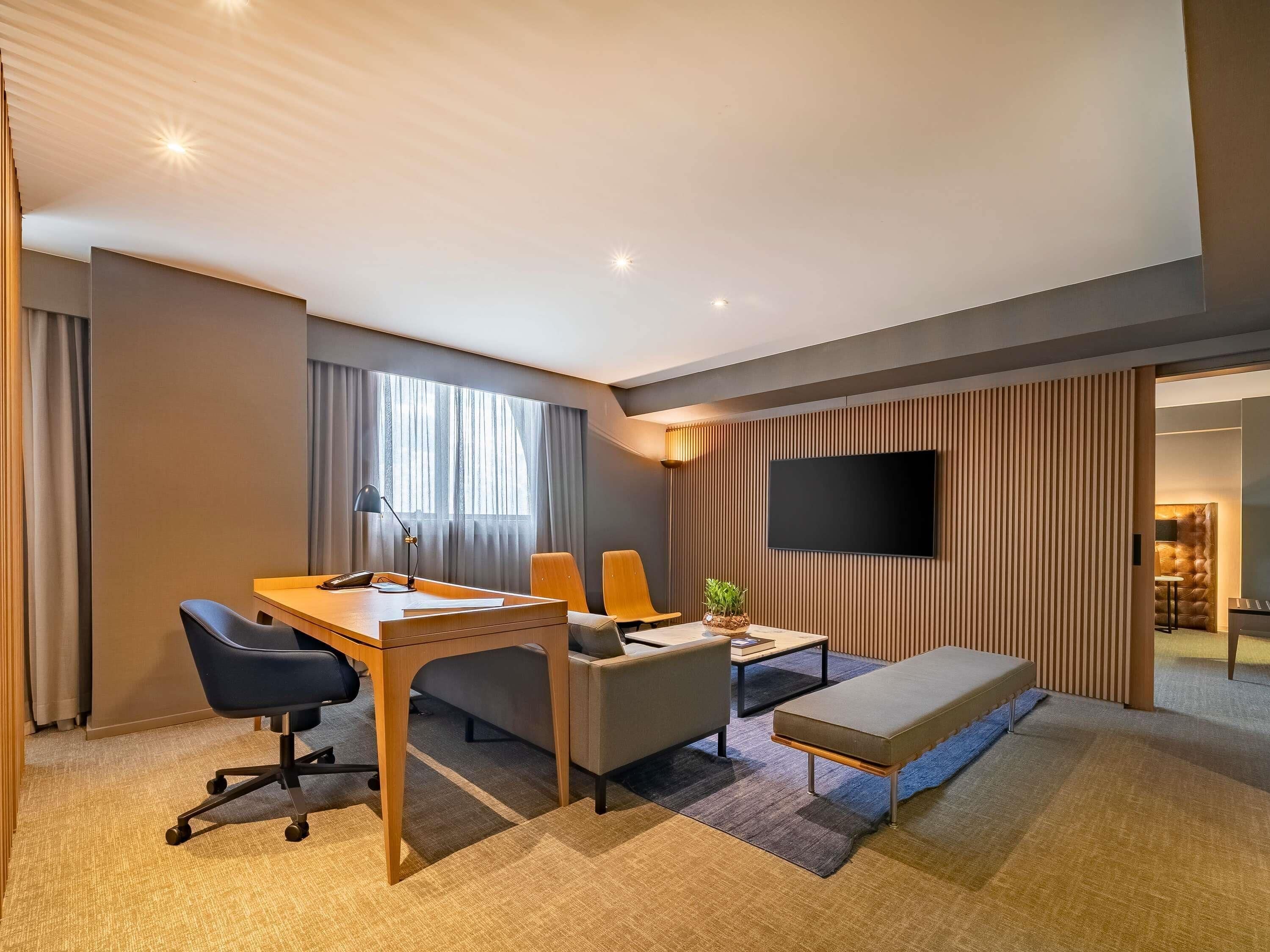 A modern hotel room with a desk, two chairs, a sofa, a TV, and a large window with curtains.