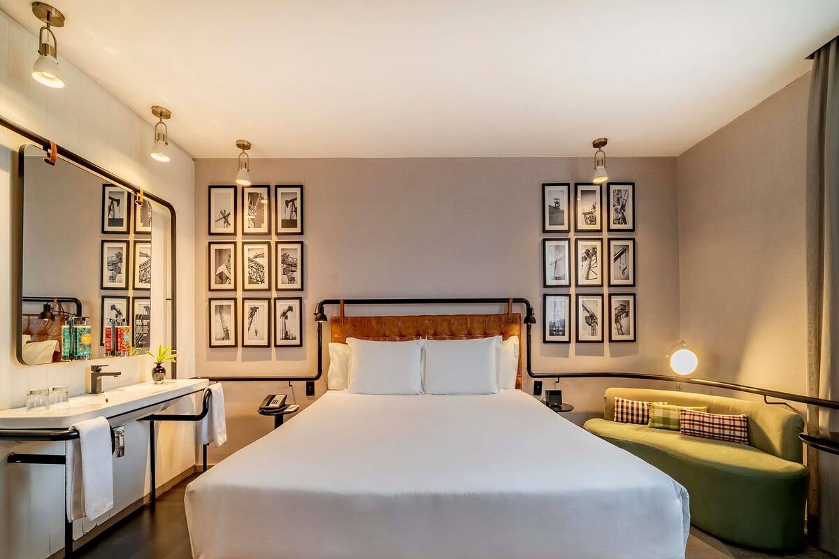 Una habitación de hotel con una cama grande, una mesita de noche con una lámpara, un sillón verde y cuadros enmarcados en las paredes.