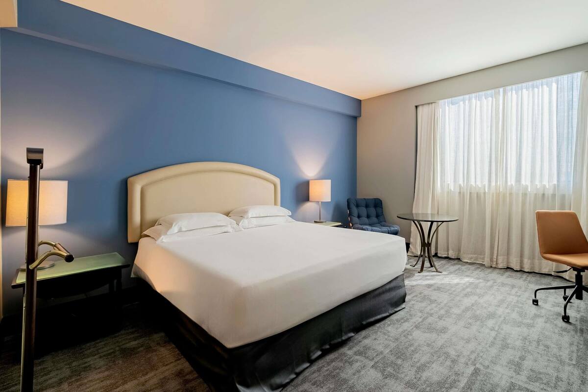 Habitación de hotel con cama, dos mesitas de noche, una silla, un escritorio y una ventana con cortinas.