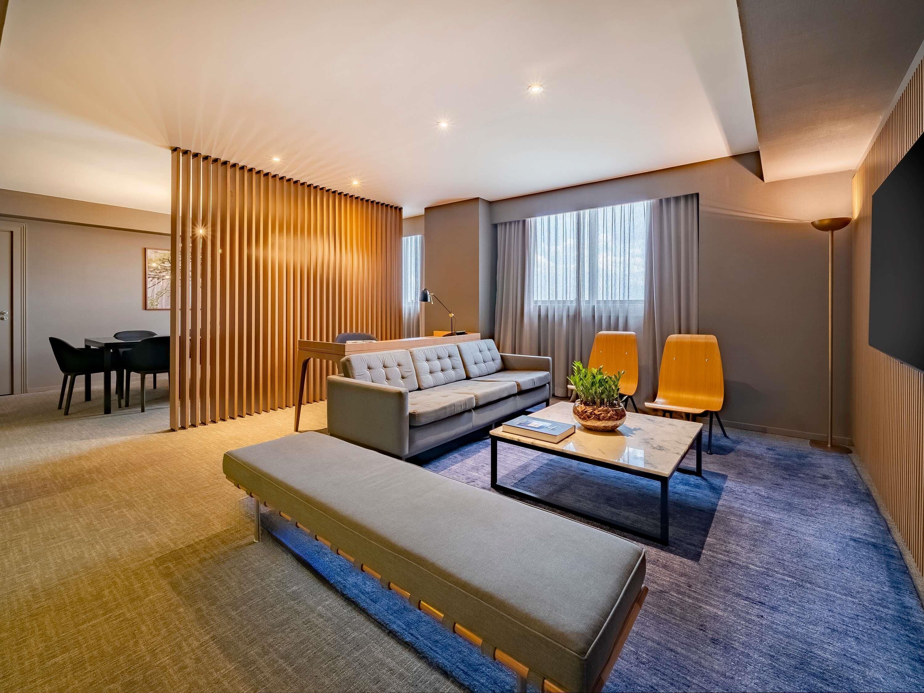 A modern hotel room with a sofa, a coffee table, a dining area, and a large window.