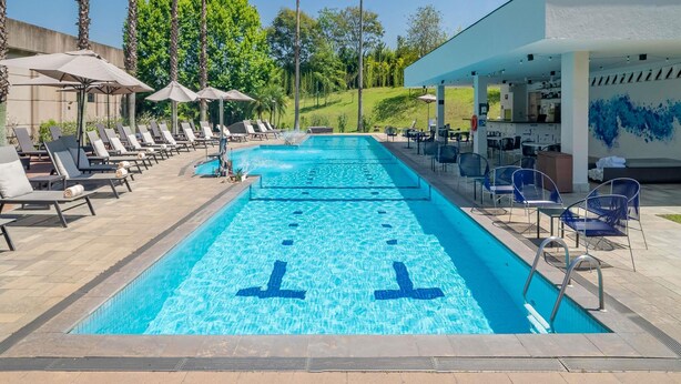 A swimming pool with lounge chairs and umbrellas, surrounded by palm trees and a covered area with tables and chairs. - Pullman São Paulo Guarulhos Airport (Guarulhos)