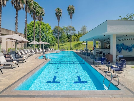 A swimming pool with lounge chairs and umbrellas, surrounded by palm trees and a covered area with tables and chairs.