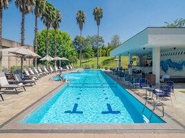 Una piscina con sillas de descanso y sombrillas, rodeada de palmeras y una zona cubierta con mesas y sillas.