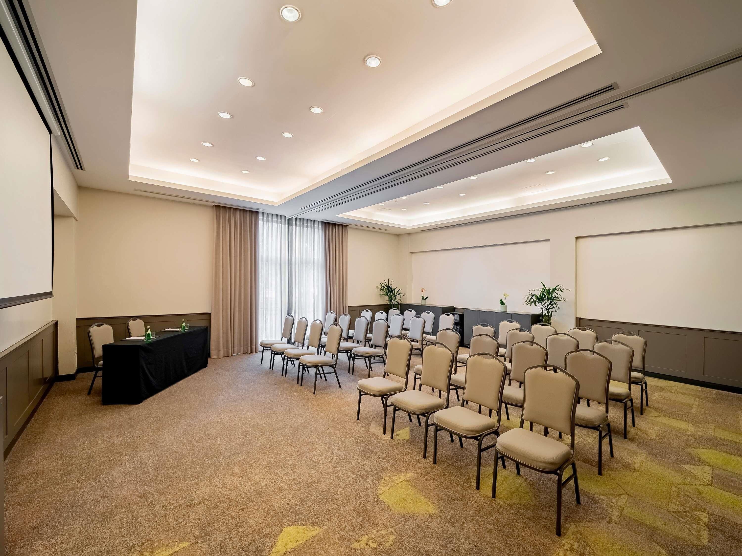 A conference room with a long table, rows of chairs, a projector screen, and a carpeted floor.