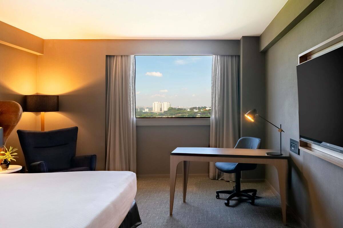 Habitación de hotel con cama, escritorio, silla, lámpara y vista del paisaje urbano a través del ventanal.