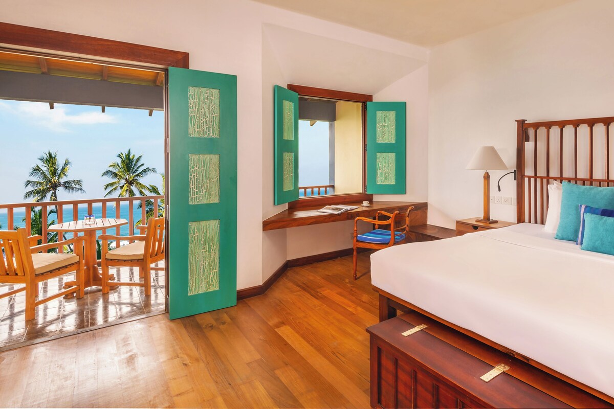 A hotel room with a balcony, wooden furniture, a bed with blue pillows, and a desk with a lamp.