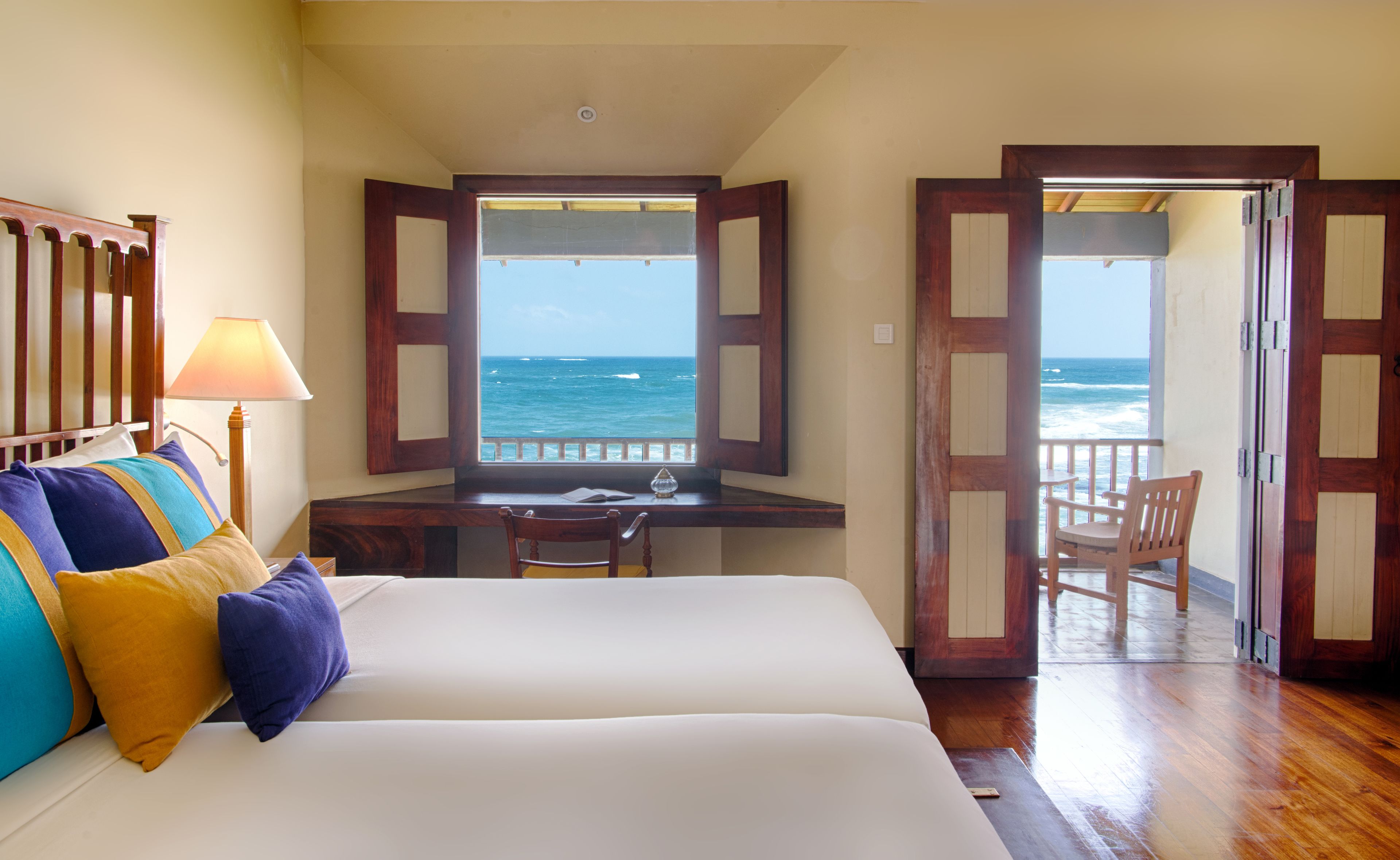 A bedroom with a bed, a desk, and a chair, a balcony with a view of the ocean, and wooden doors.