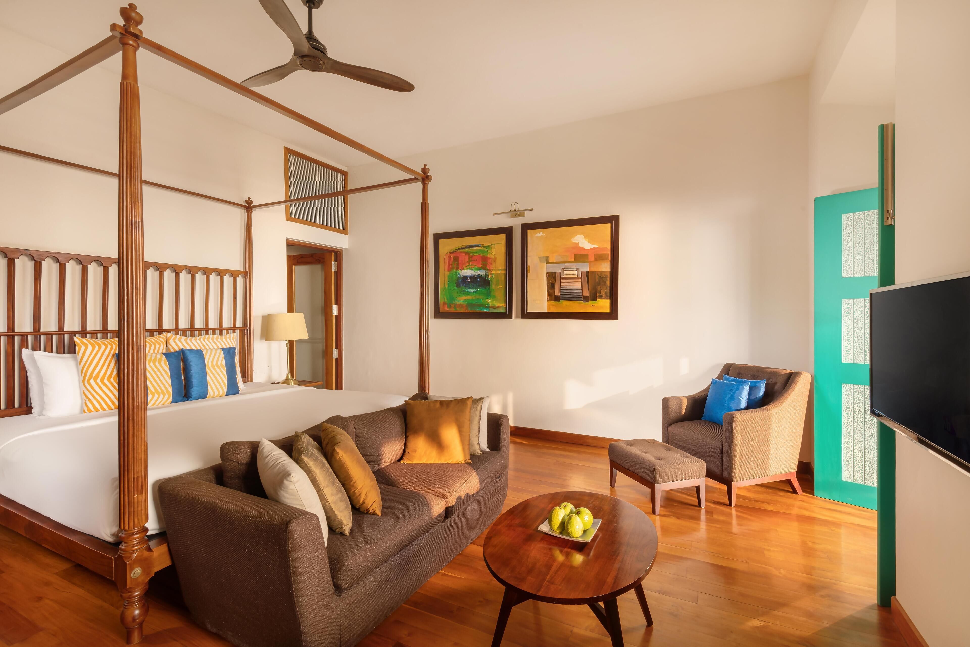 A four-poster bed with a wooden canopy, a sofa, a small round table with fruit, a chair, and a television.