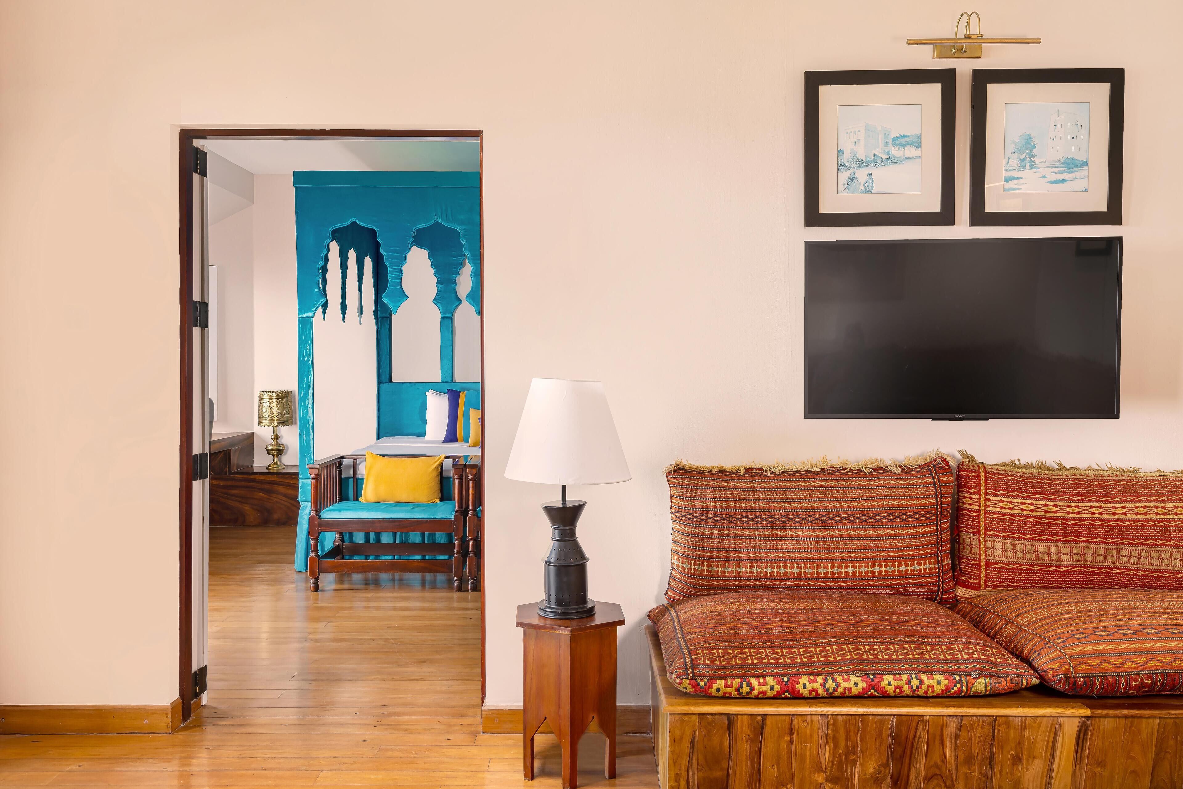 A living room with a wooden floor, a sofa with patterned cushions, a lamp, and a TV mounted on the wall.