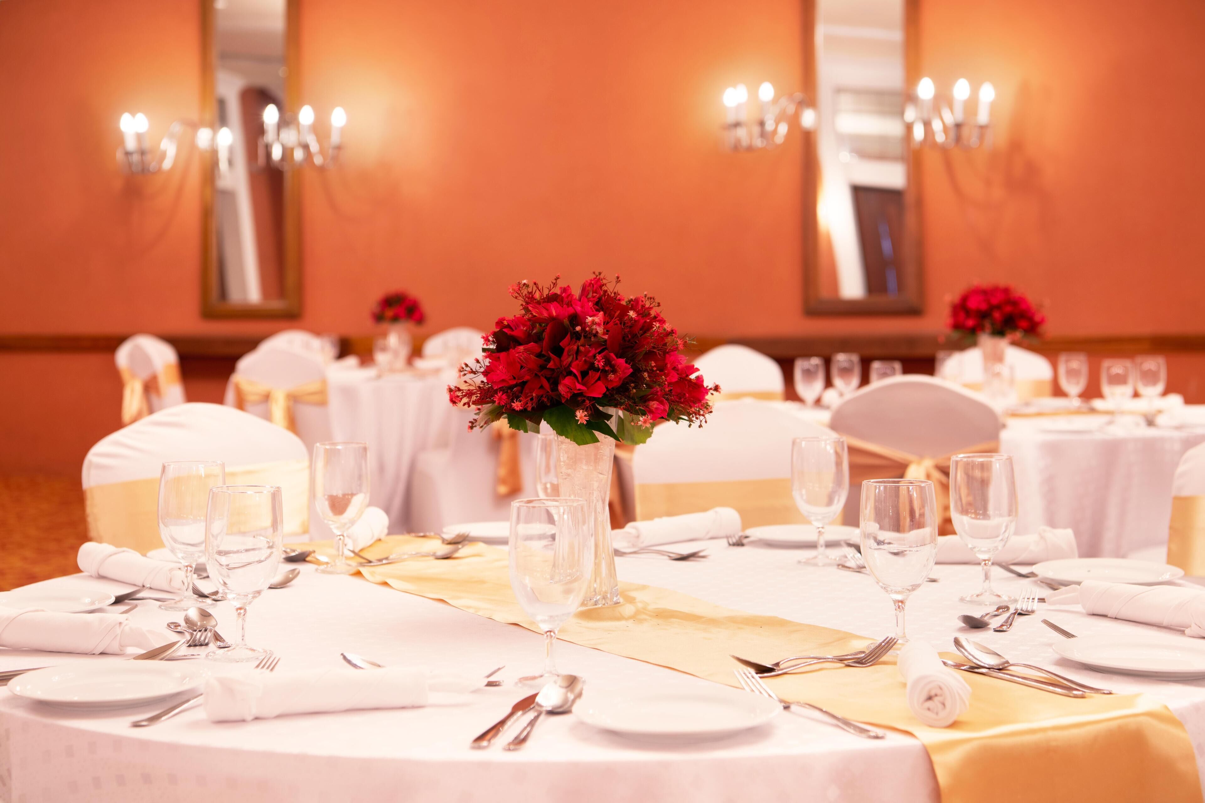 Une salle de banquet avec des tables rondes dressées pour un événement formel, agrémentée de nappes blanches, de rubans dorés et de compositions florales rouges.