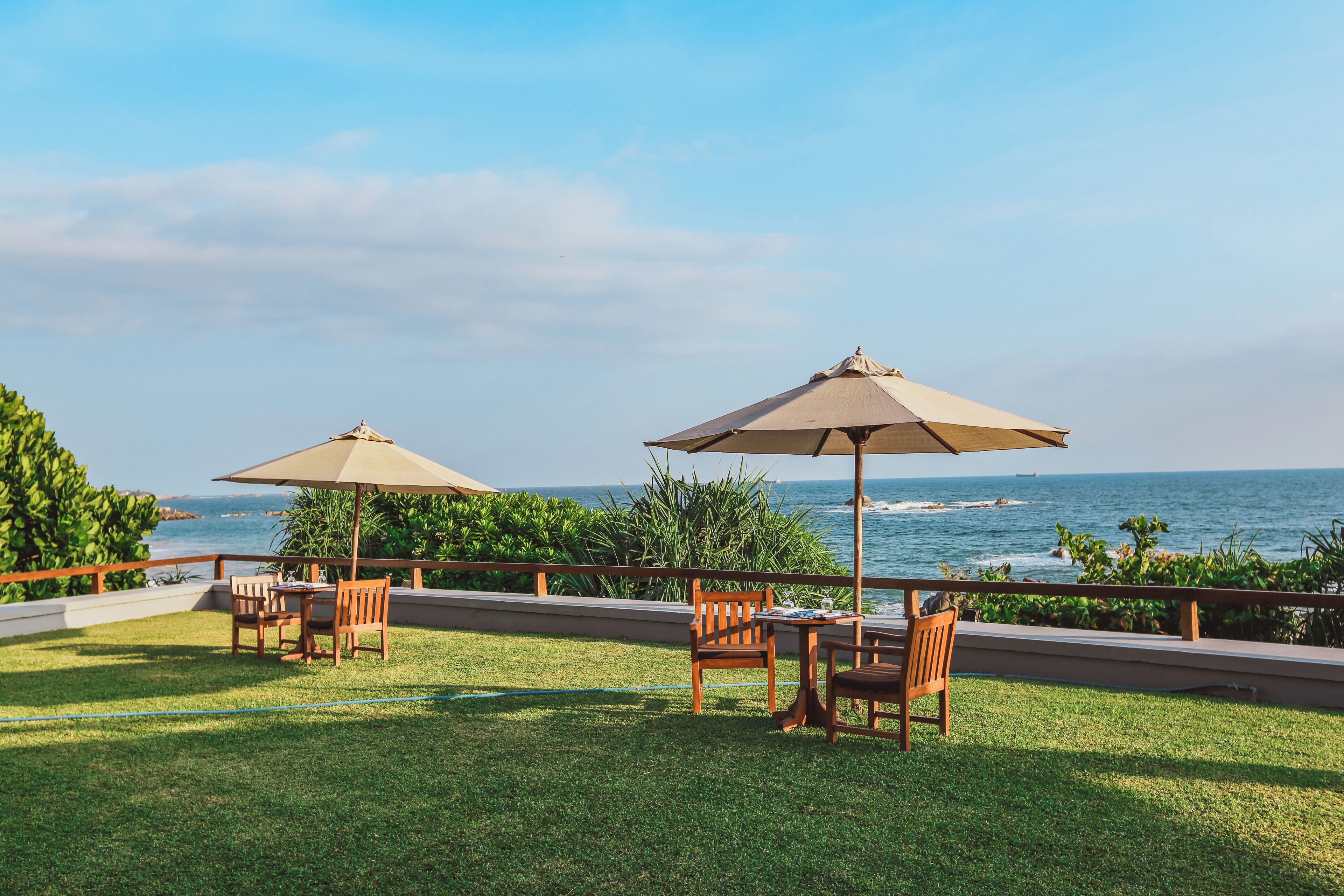 An outdoor seating area with two wooden tables and chairs, each under a beige umbrella, overlooking the ocean.