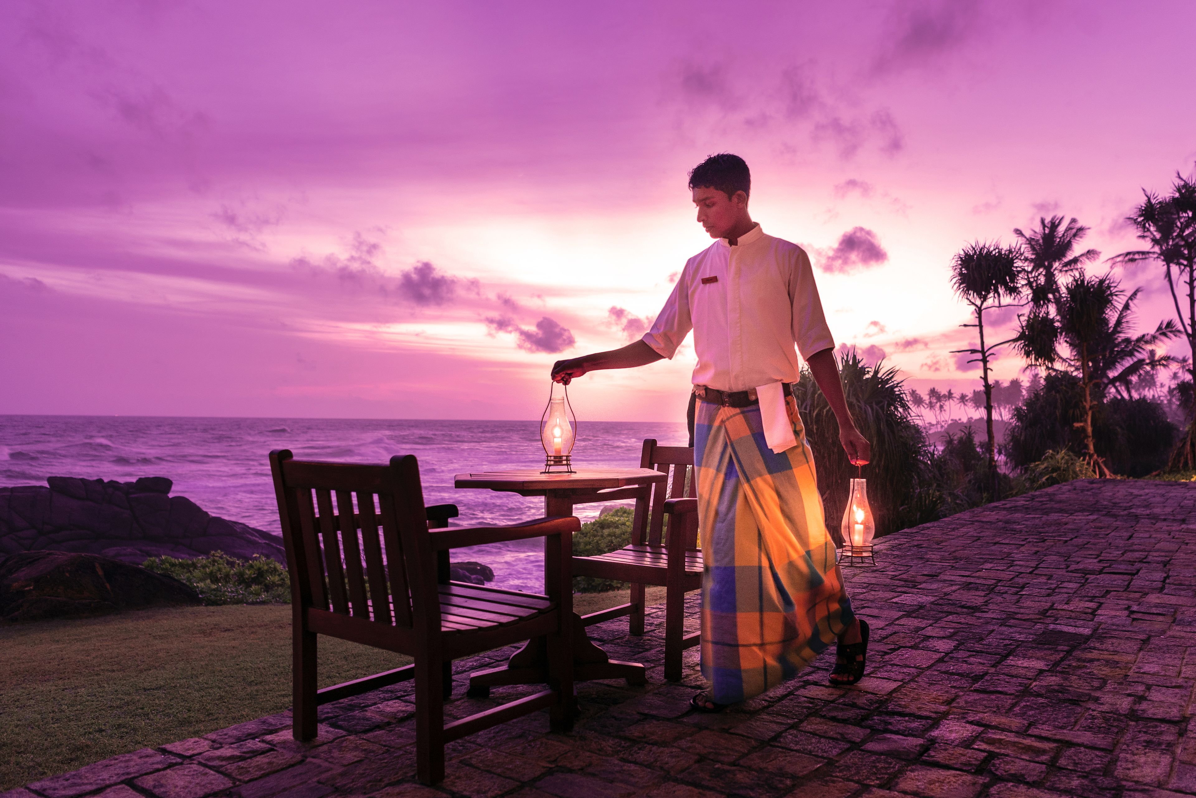 Un hombre con atuendo tradicional está de pie junto a una mesa con sillas, con vista a una costa rocosa y el océano al fondo.