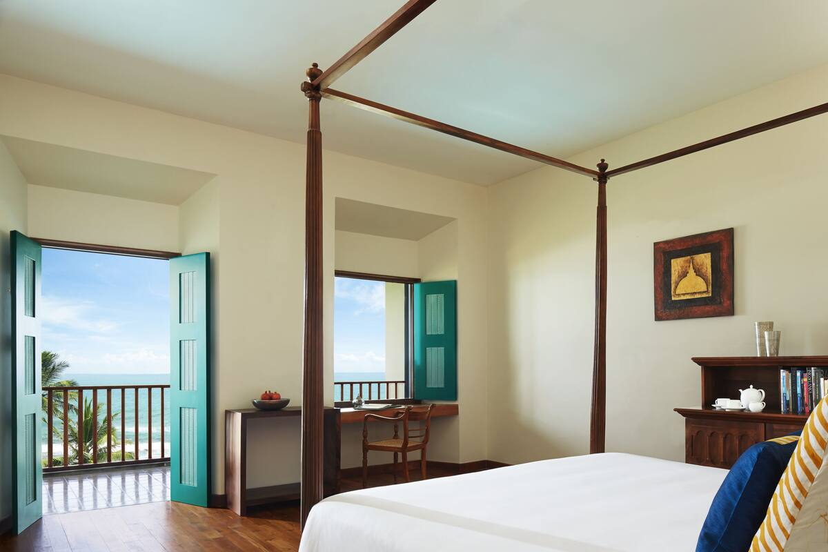 A room with a four-poster bed, a desk with a chair, and a view of the beach.