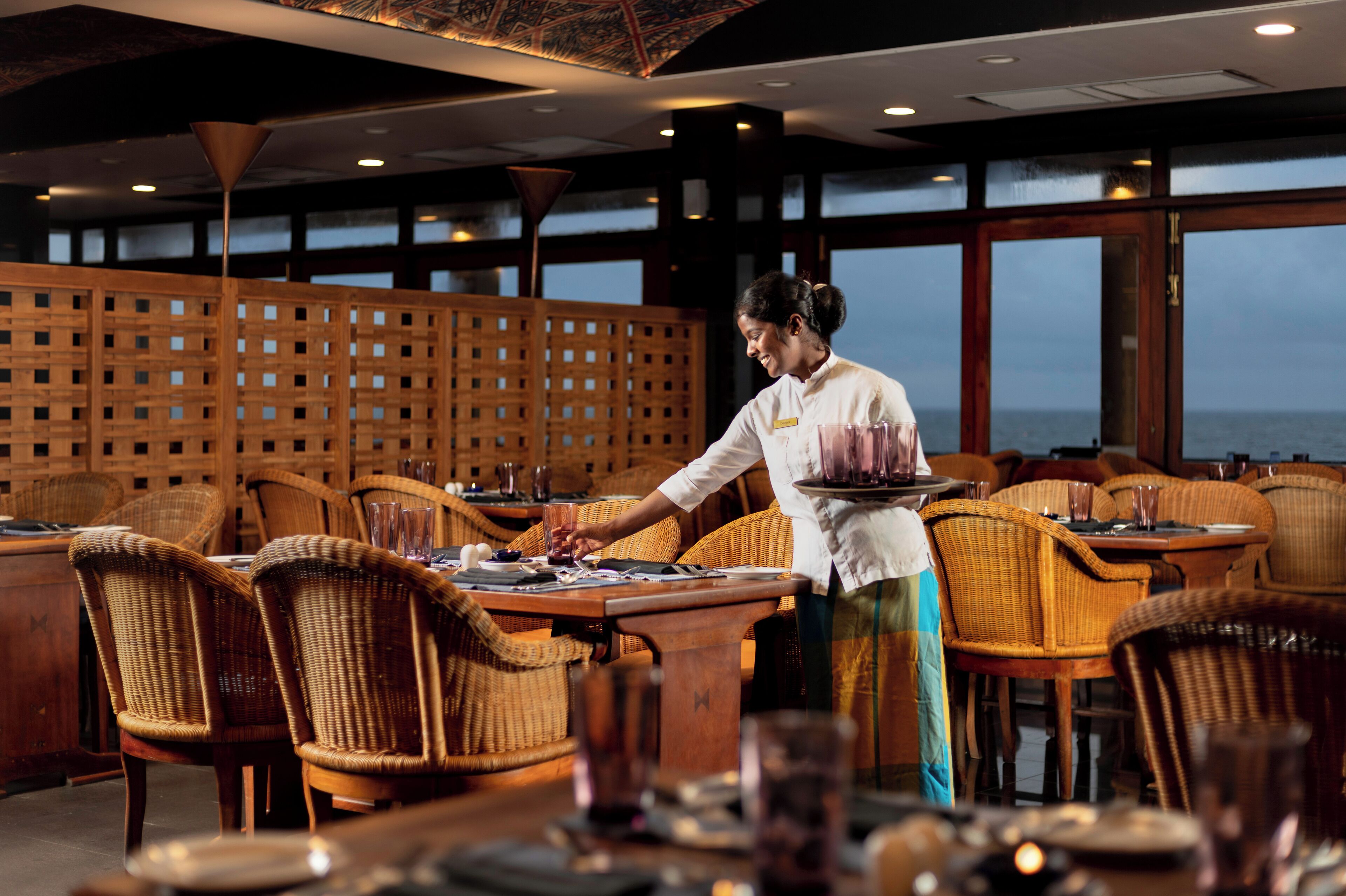 A restaurant with wicker chairs and tables, a wooden partition, and a view of the sea.