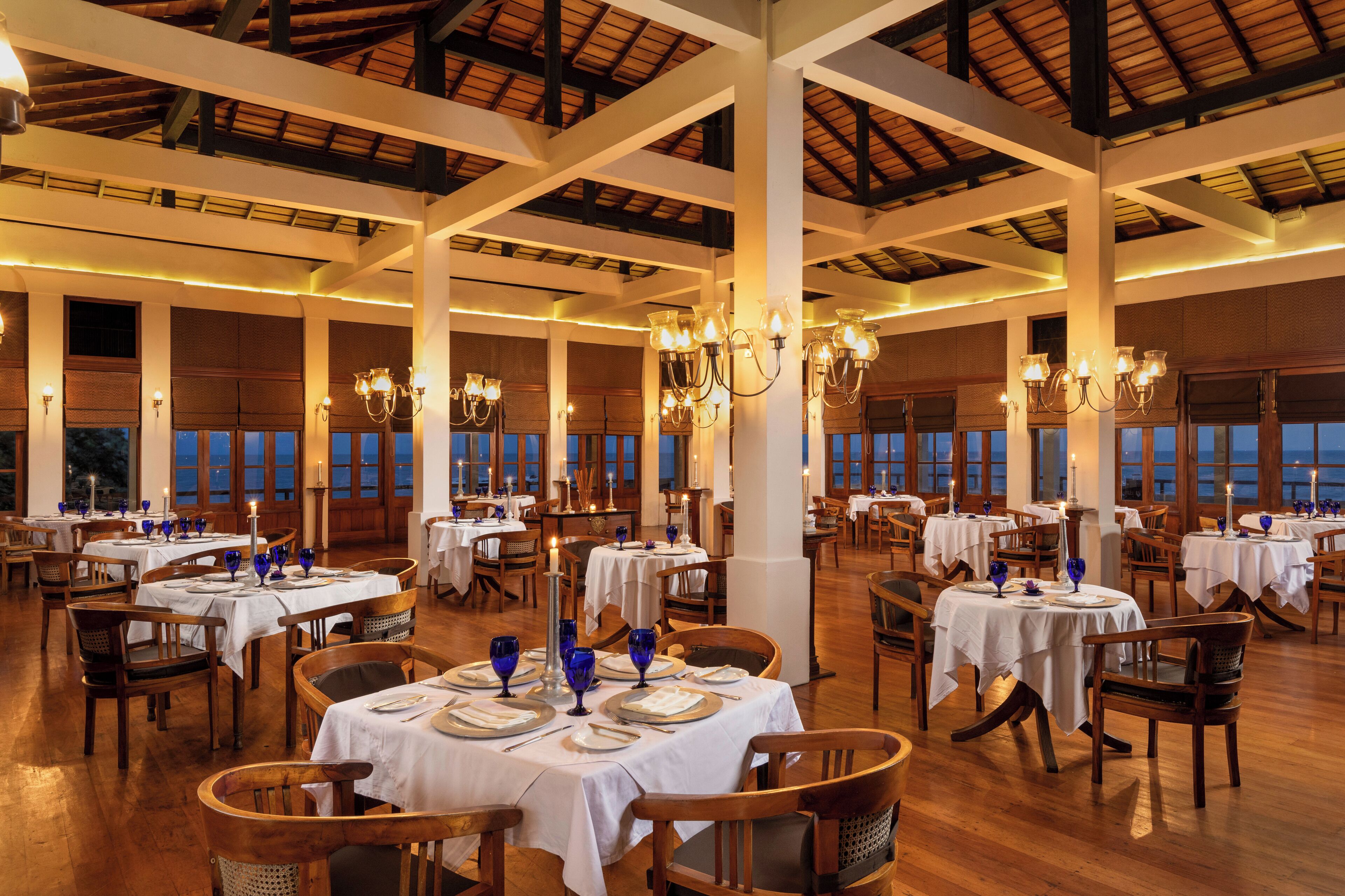 A spacious dining area with multiple tables set for dining, large windows, and a high ceiling with exposed beams.