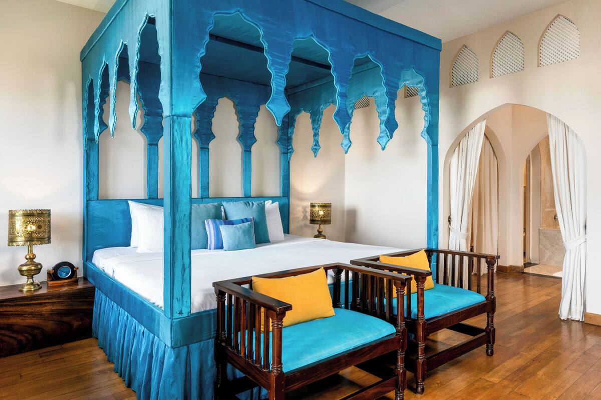 A four-poster bed with a blue canopy, two wooden chairs with cushions, a bedside table with a lamp, and a mirror.