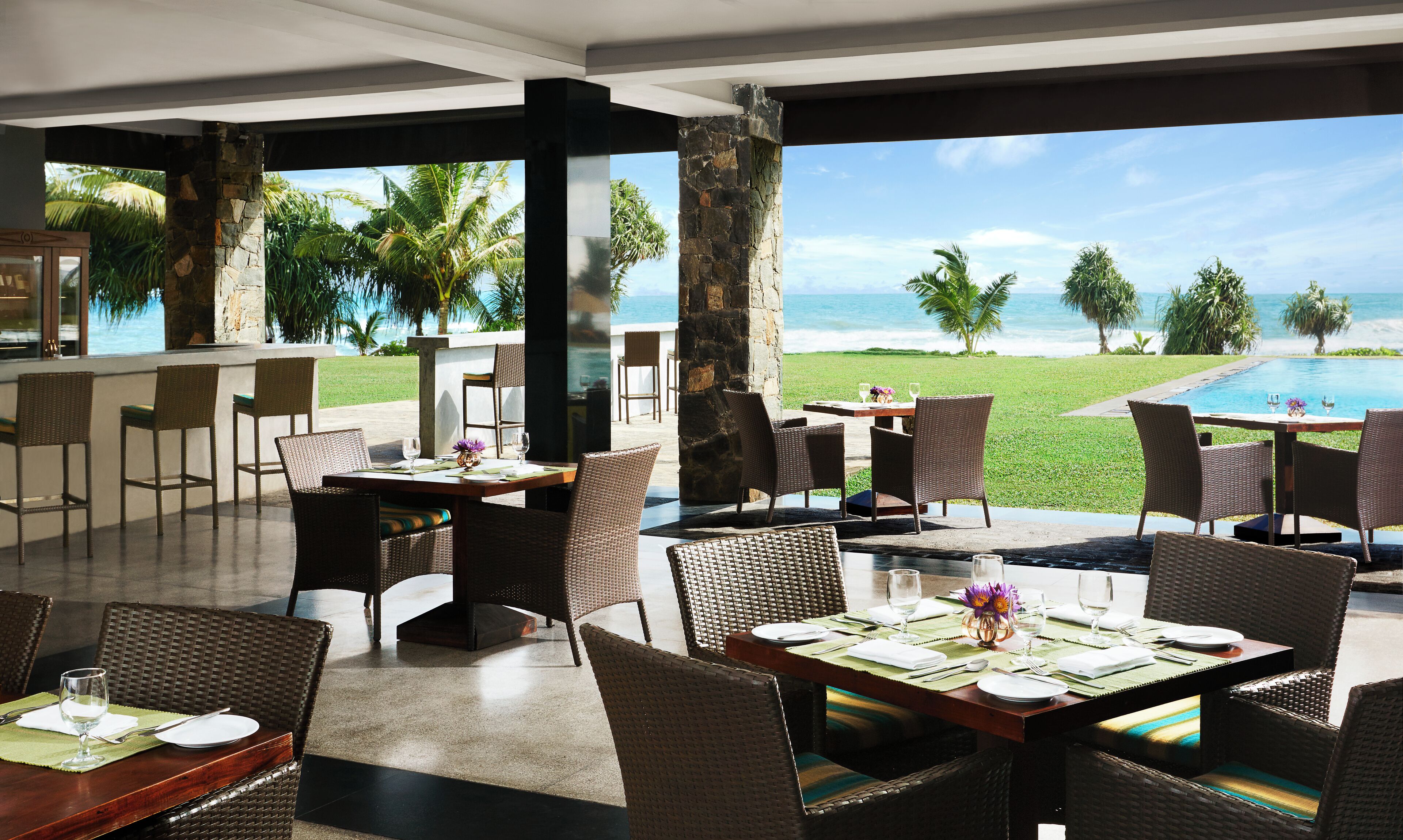 A restaurant with outdoor seating overlooking a pool and beach, featuring wicker furniture and a clear view of the ocean.