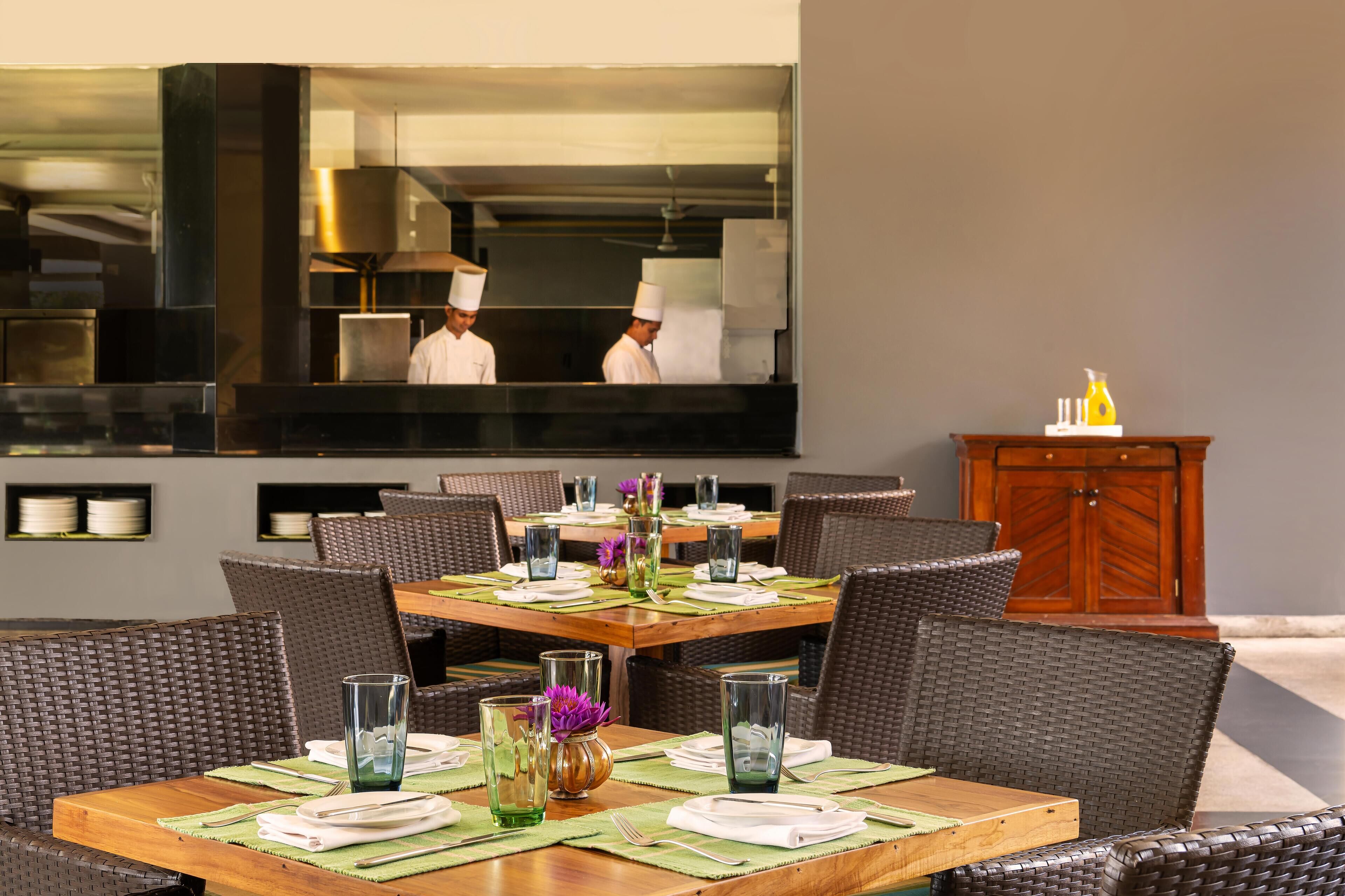 A restaurant dining area with wicker chairs, wooden tables, and a large mirror reflecting the kitchen and chefs.