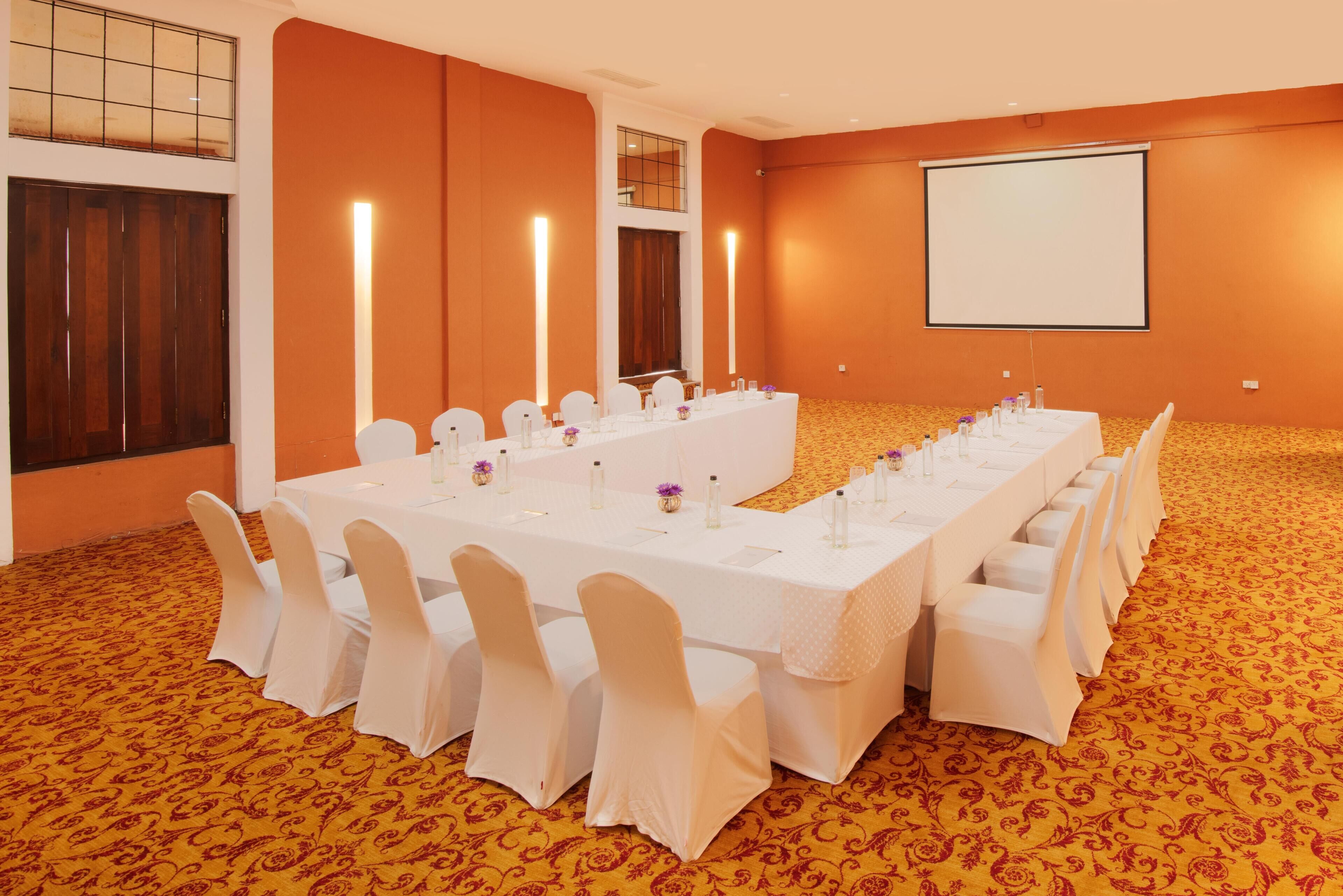 Uma sala de conferência com uma mesa comprida, cadeiras brancas e uma tela de projetor na parede.