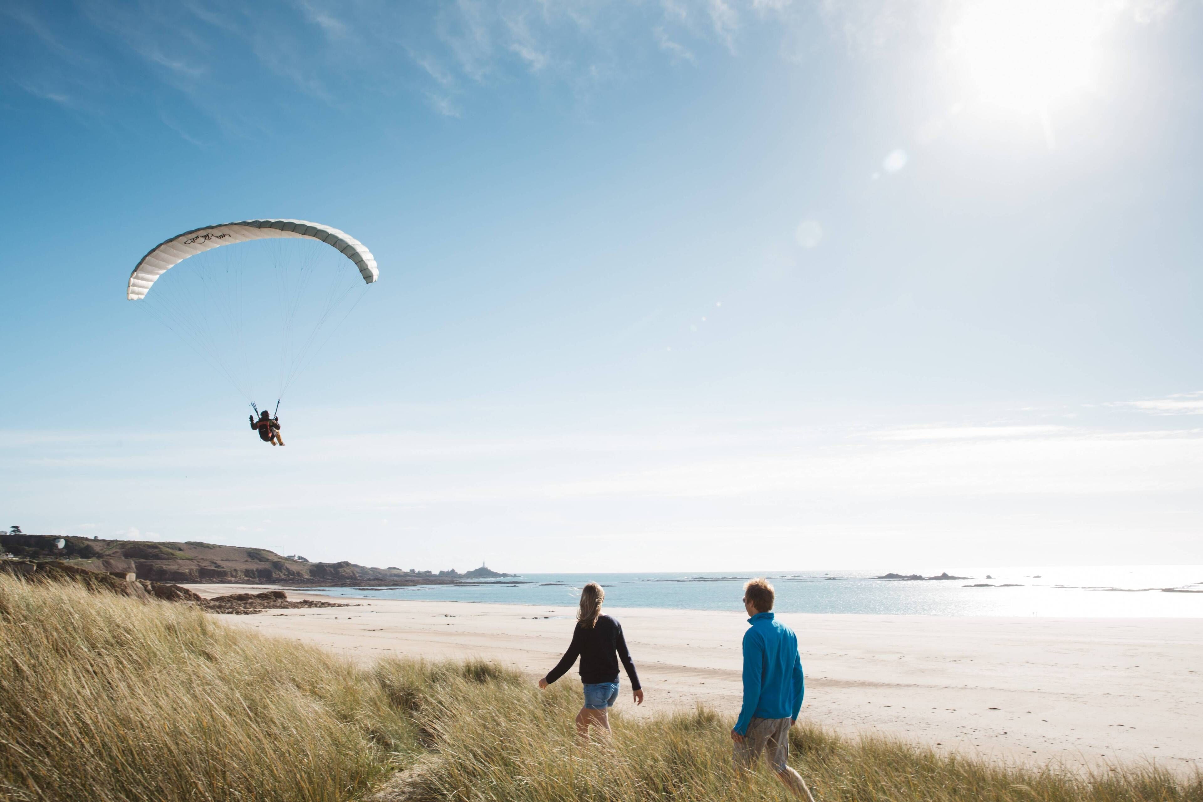 A beach with a person paragliding in the background and two people walking on the sand.
