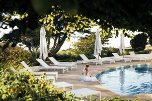 Zona junto a la piscina con sillones de descanso y sombrillas, rodeada de vegetación.