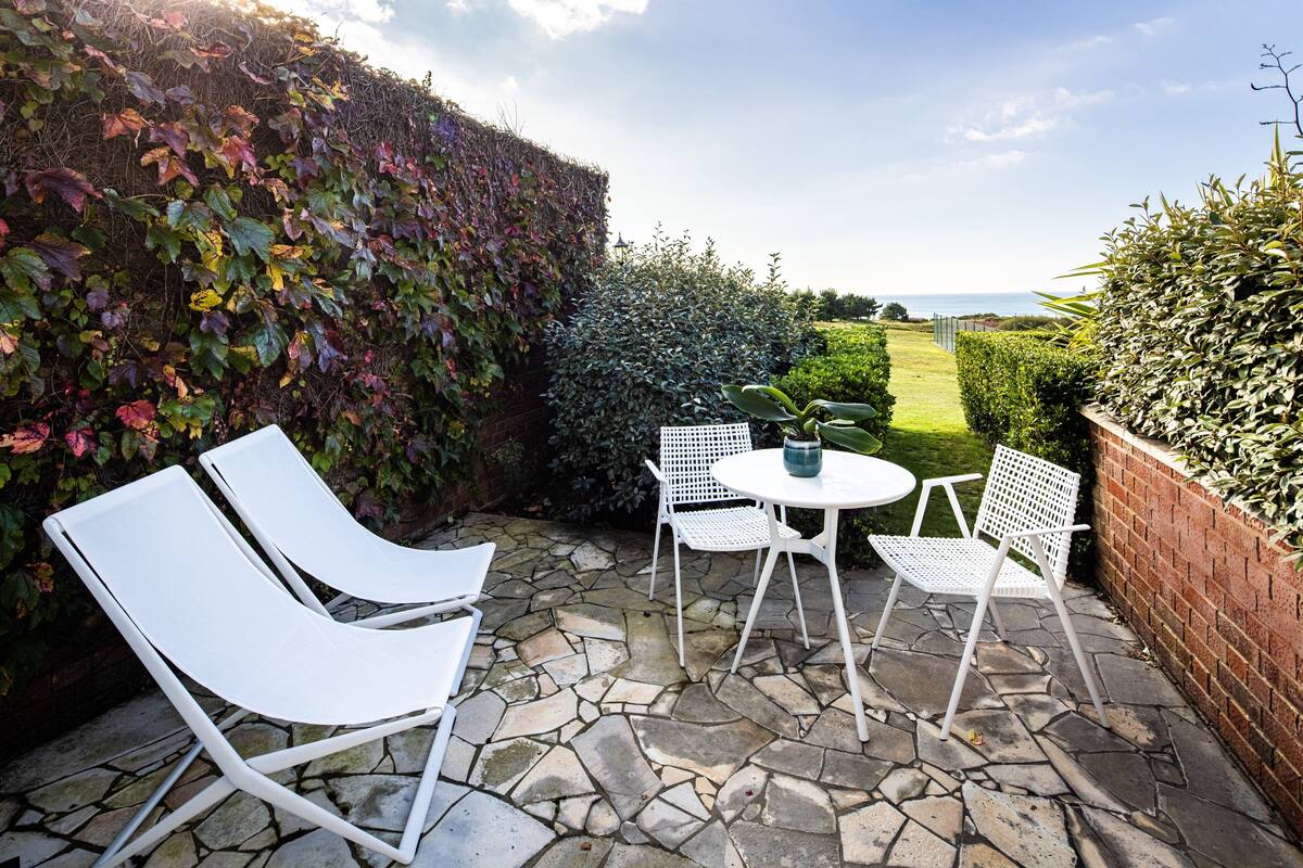 Un patio con sillas blancas y una mesa, piso de piedra y vista al mar.