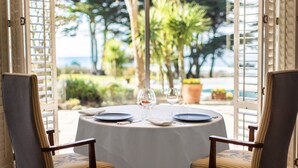 A round table with a white tablecloth, two chairs with patterned upholstery, and a view of a garden and ocean through open white shutters.