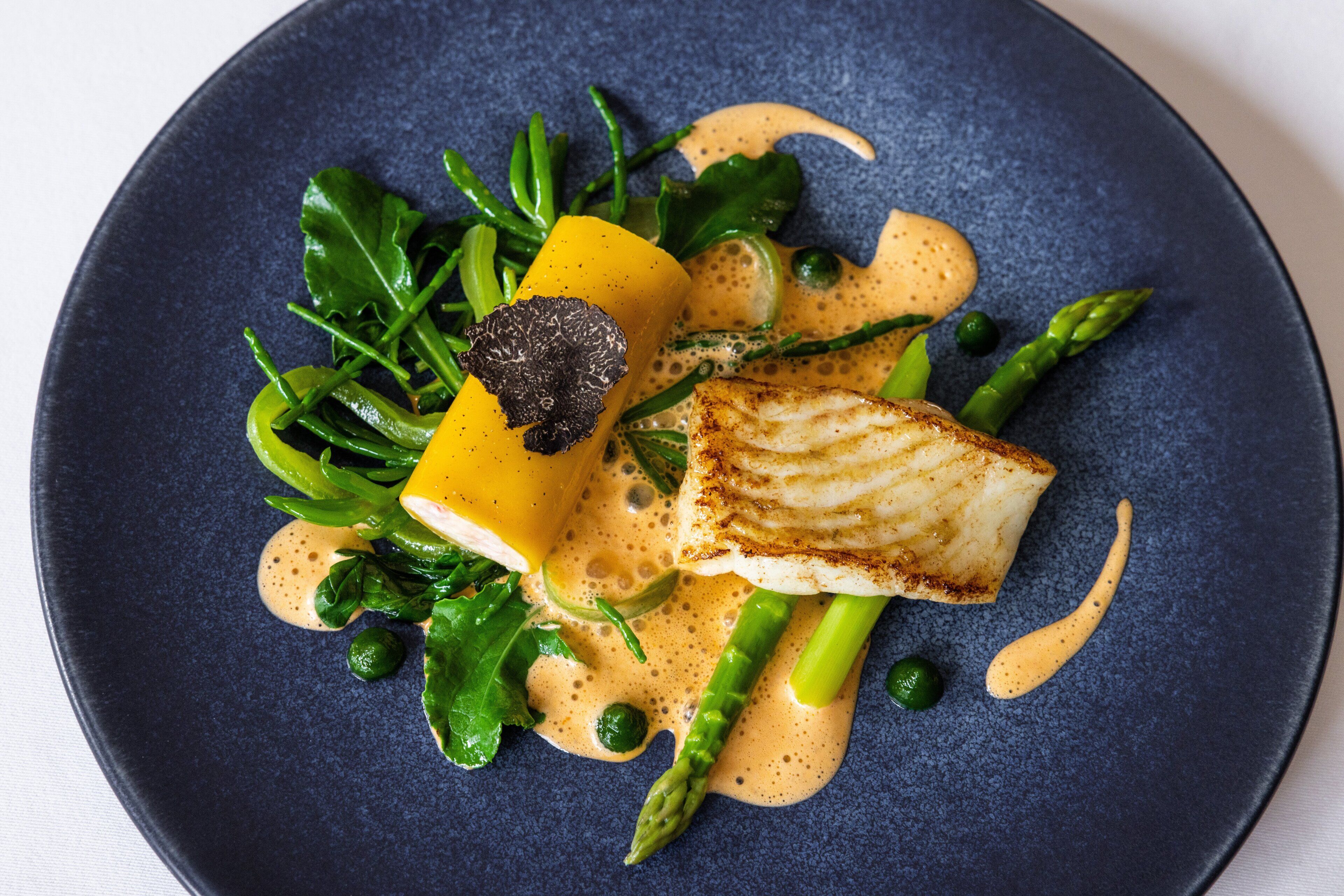Une assiette bleu foncé contenant un morceau de poisson, des asperges et des légumes verts.