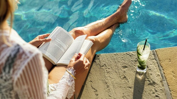 A person relaxing by a poolside with a book and a drink.