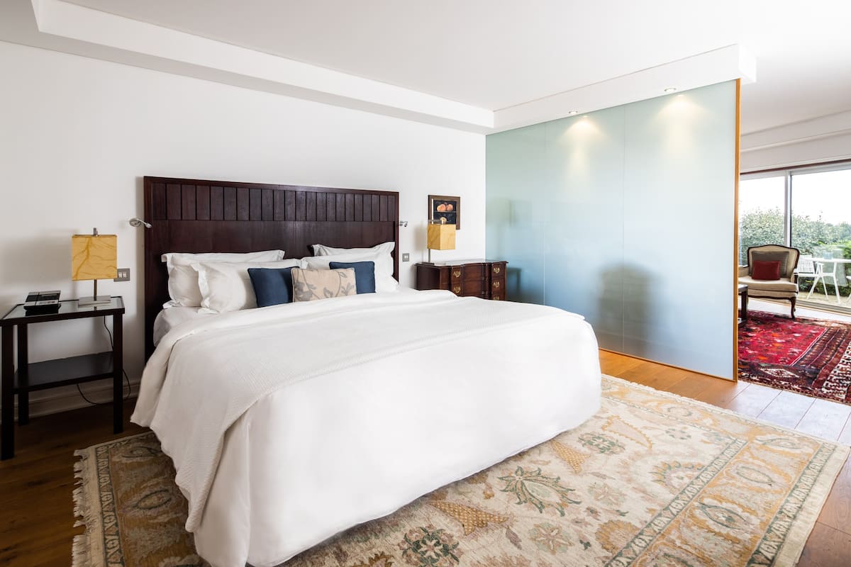 Un dormitorio moderno con una cama grande, una mesita de noche, una lámpara, un cabecero de madera y vistas al exterior a través de un ventanal.