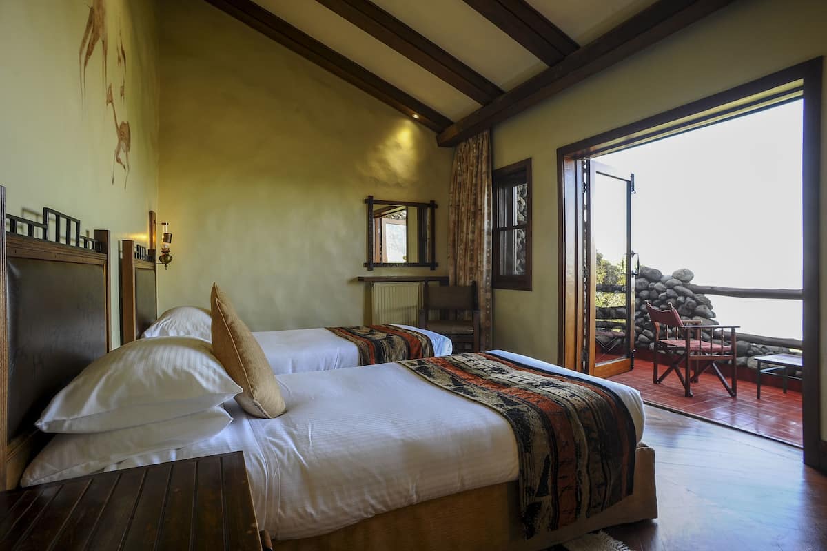 A bedroom with two beds, a wooden ceiling, a large window, and a view of a stone wall outside.
