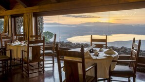 Uma sala de jantar com mesas e cadeiras, vista para um lago e montanhas, e uma parede de pedra.