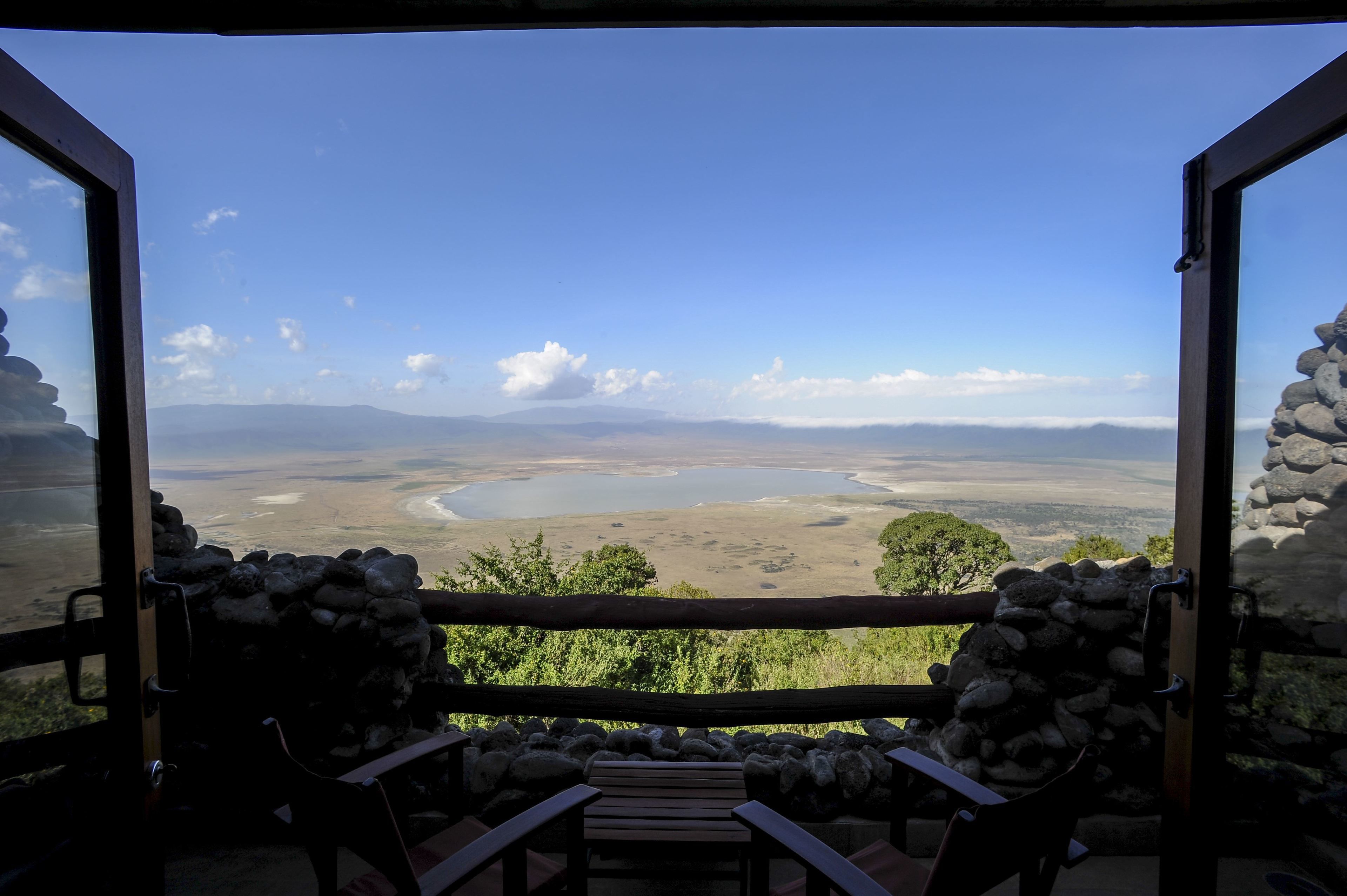 A view from a balcony with stone walls and wooden furniture, overlooking a valley with a river and distant mountains.