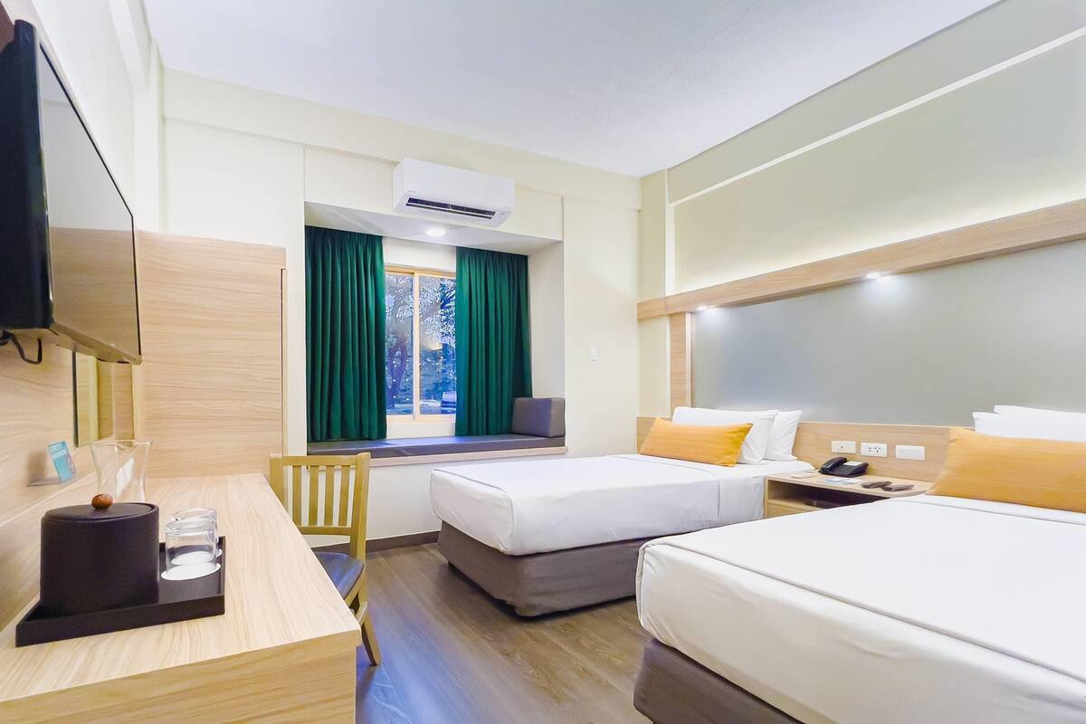 A hotel room with a desk, two beds, a window with curtains, an air conditioner, and a bedside table with a phone.