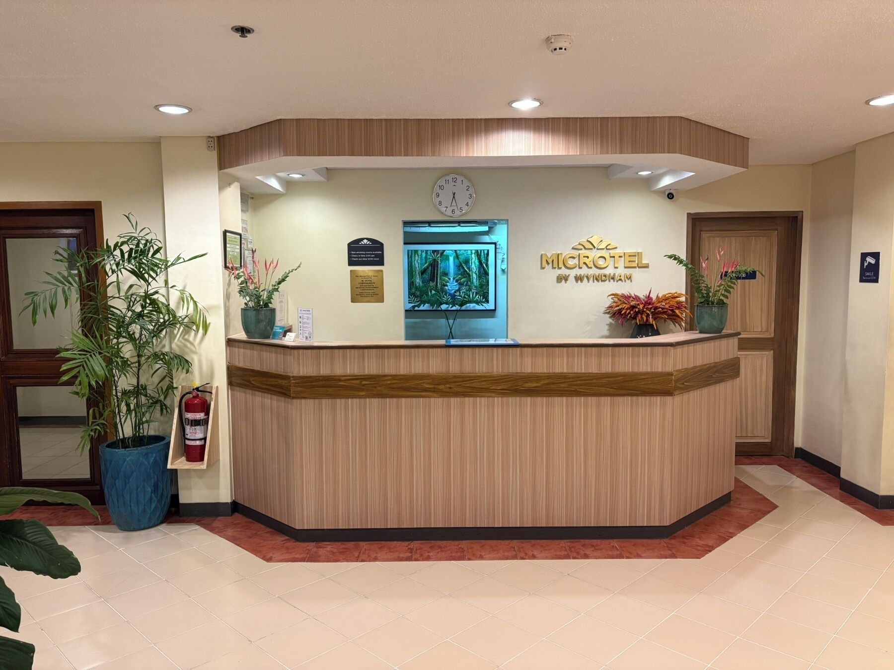 En receptionsdisk med en klocka, en skylt med texten ”MICROTEL BY WYNDHAM” och en brandsläckare. Det finns krukväxter och en inramad tavla på väggen.