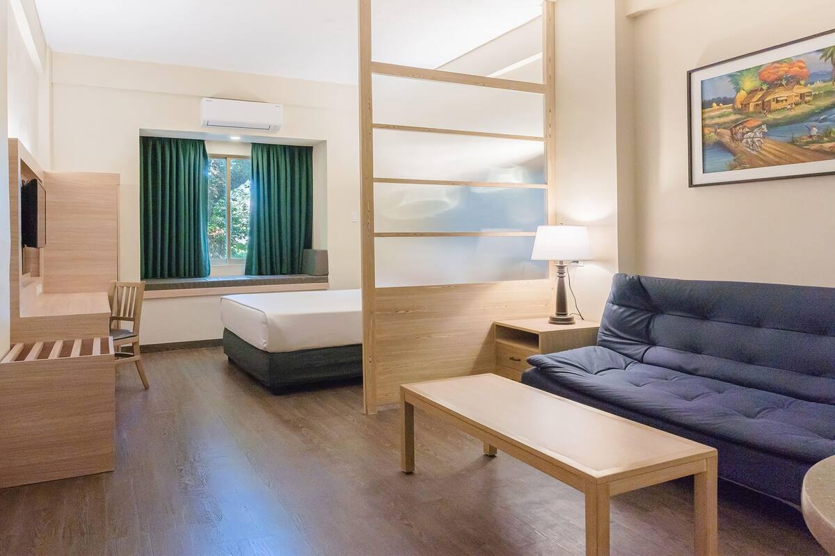 A hotel room with a bed, a sofa, a desk, a chair, a window with curtains, a lamp, and a painting on the wall.