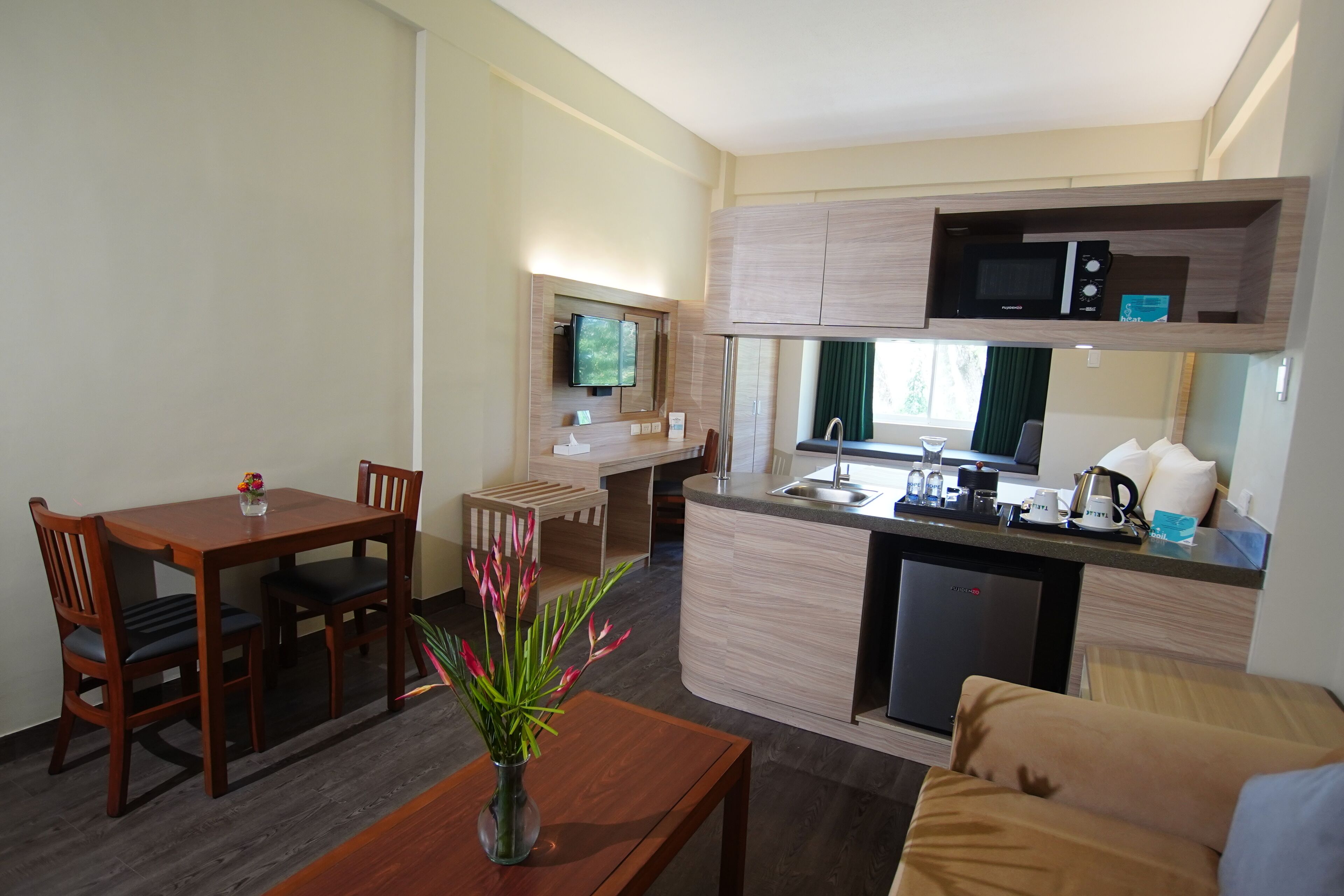 A compact hotel room with a wooden dining table, a small kitchen area with a microwave, and a sofa.