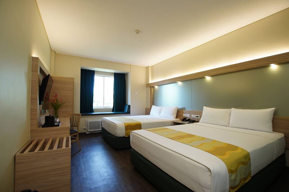 A hotel room with two beds, a desk, a chair, a window with curtains, and a wall-mounted TV.