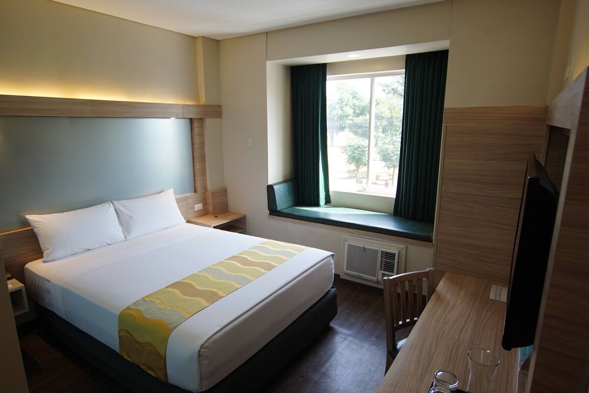 A hotel room with a large bed, a window with curtains, a desk with a chair, and a television.