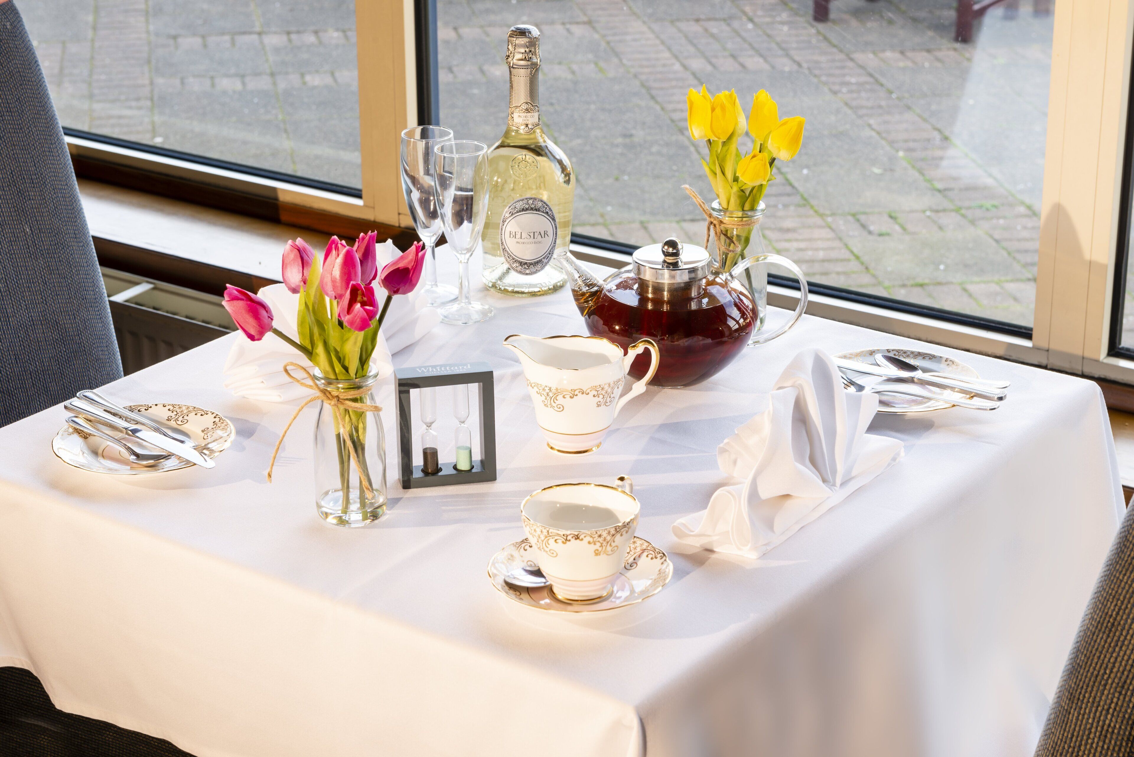 A table set for a meal with a bottle of Belstar champagne, a teapot, and a vase with yellow flowers.