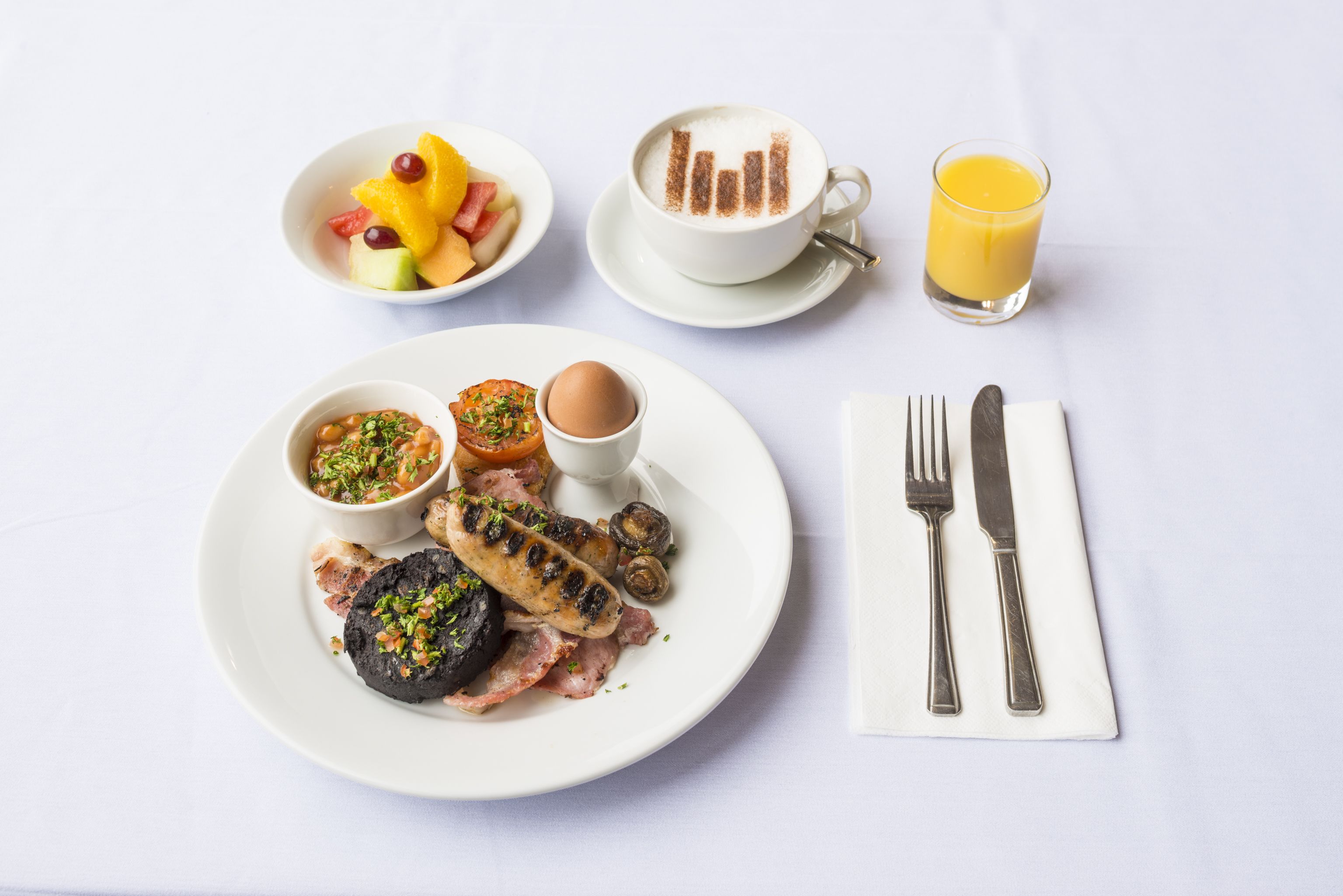 A breakfast setting with a plate of food, a cup of coffee, a glass of orange juice, and cutlery on a white tablecloth.
