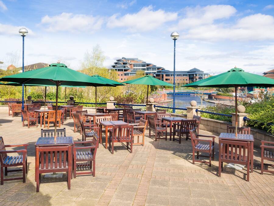 An outdoor patio area with wooden tables and chairs, green umbrellas, and a view of buildings in the background.