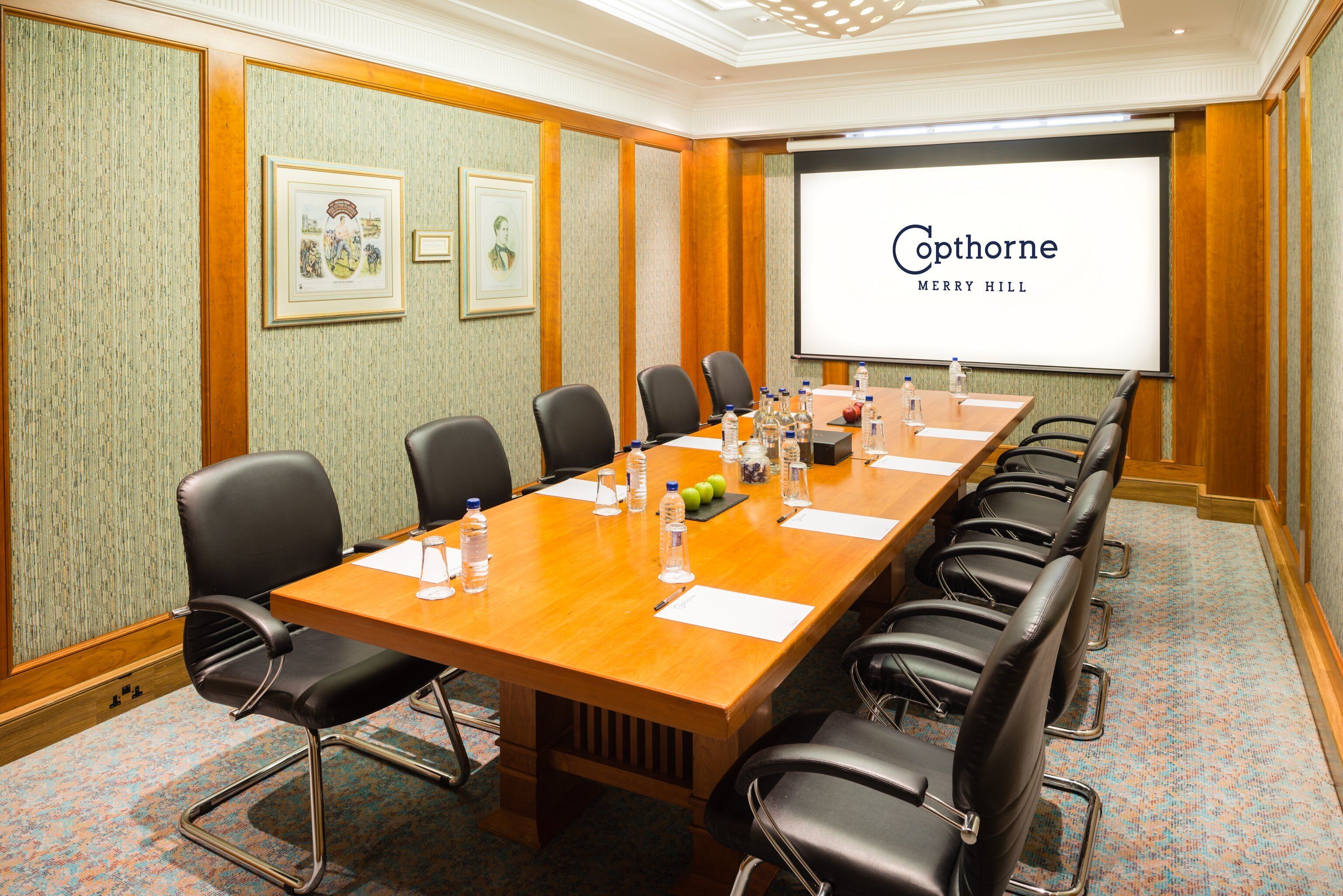 A conference room with a long table, black chairs, and a projector screen displaying "Cophorne Merry Hill".