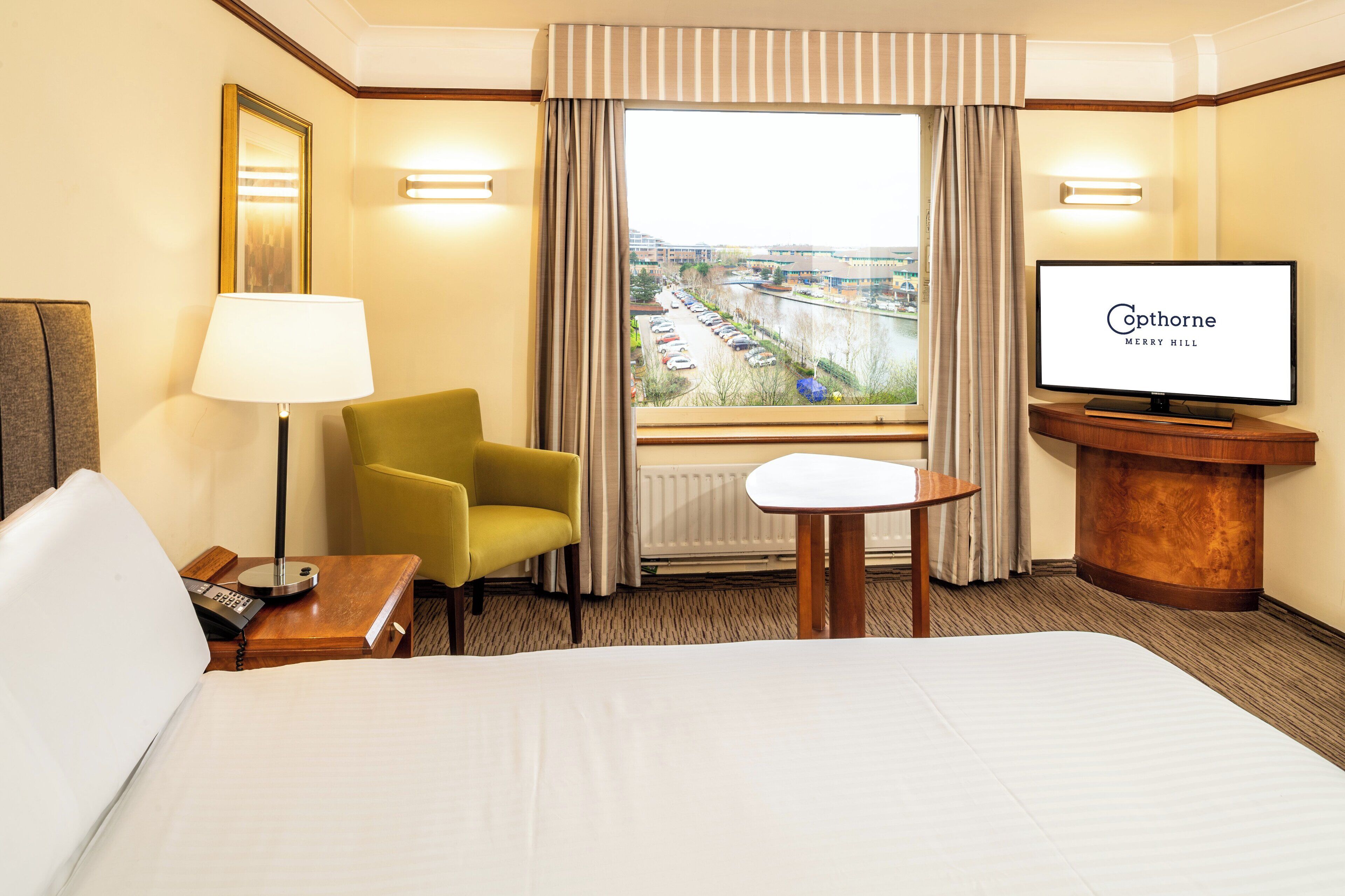 Habitación de hotel con cama, escritorio con lámpara, televisión y vista a un estacionamiento y edificios.